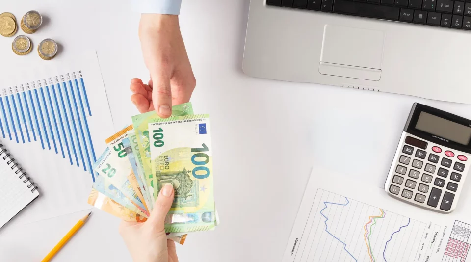 Mesa de secretaria com mão a segurar notas de euro, calculadora e computador portátil