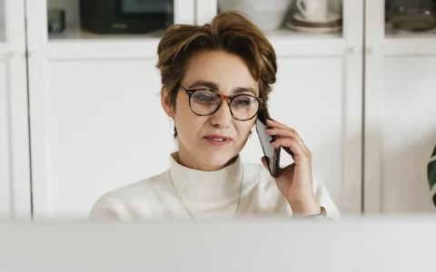 Mulher de óculos falando ao celular em frente ao computador em um ambiente iluminado