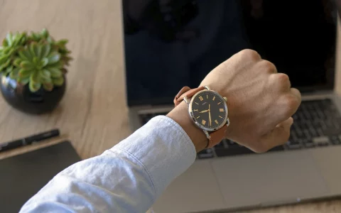 Close de uma pessoa olhando o relógio de pulso com um laptop ao fundo