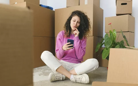 Mulher sentada no chão entre caixas de mudança, olhando para o celular com expressão pensativa