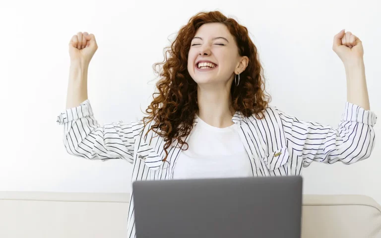 Jovem ruiva a celebrar com os punhos erguidos e um sorriso aberto em frente a um computador portátil, com fundo branco.