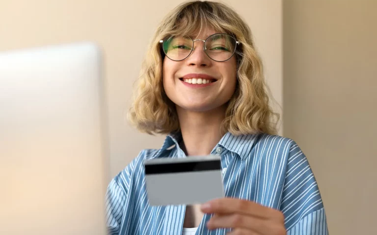 Jovem loira de óculos e camisa às riscas azuis a sorrir em frente ao portátil, segurando um cartão bancário cinzento.