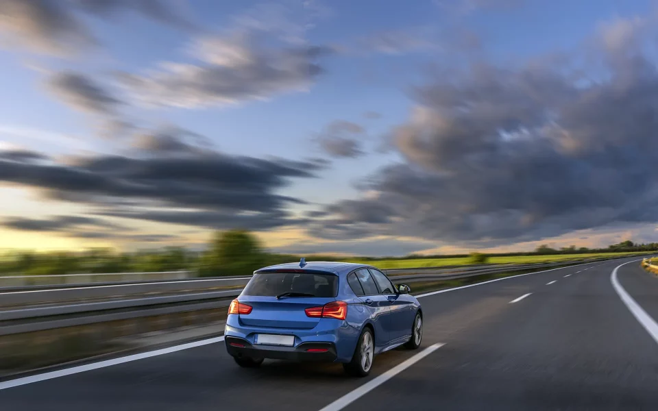 Traseira de um carro azul em movimento numa autoestrada vazia com campos verdes e um céu nublado ao fundo durante o final do dia.