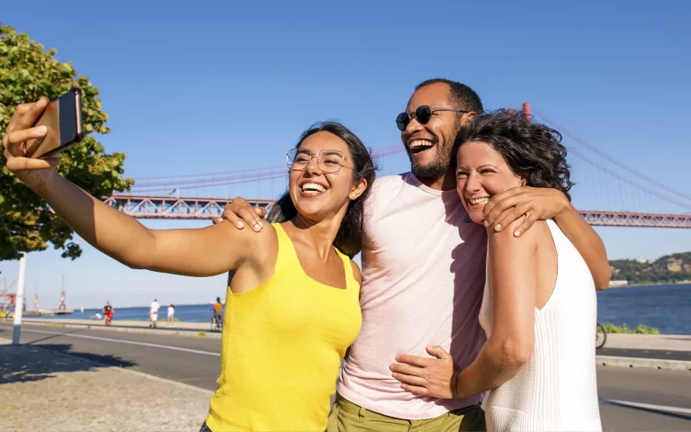 Três amigos sorridentes a tirar uma selfie junto ao rio Tejo, com a Ponte 25 de Abril em Lisboa visível ao fundo num dia de sol.