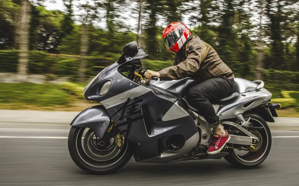 Fotografia de ação lateral de um motociclista com capacete vermelho a pilotar uma Suzuki Hayabusa cinzenta prateada em alta velocidade numa estrada ladeada por árvores desfocadas.