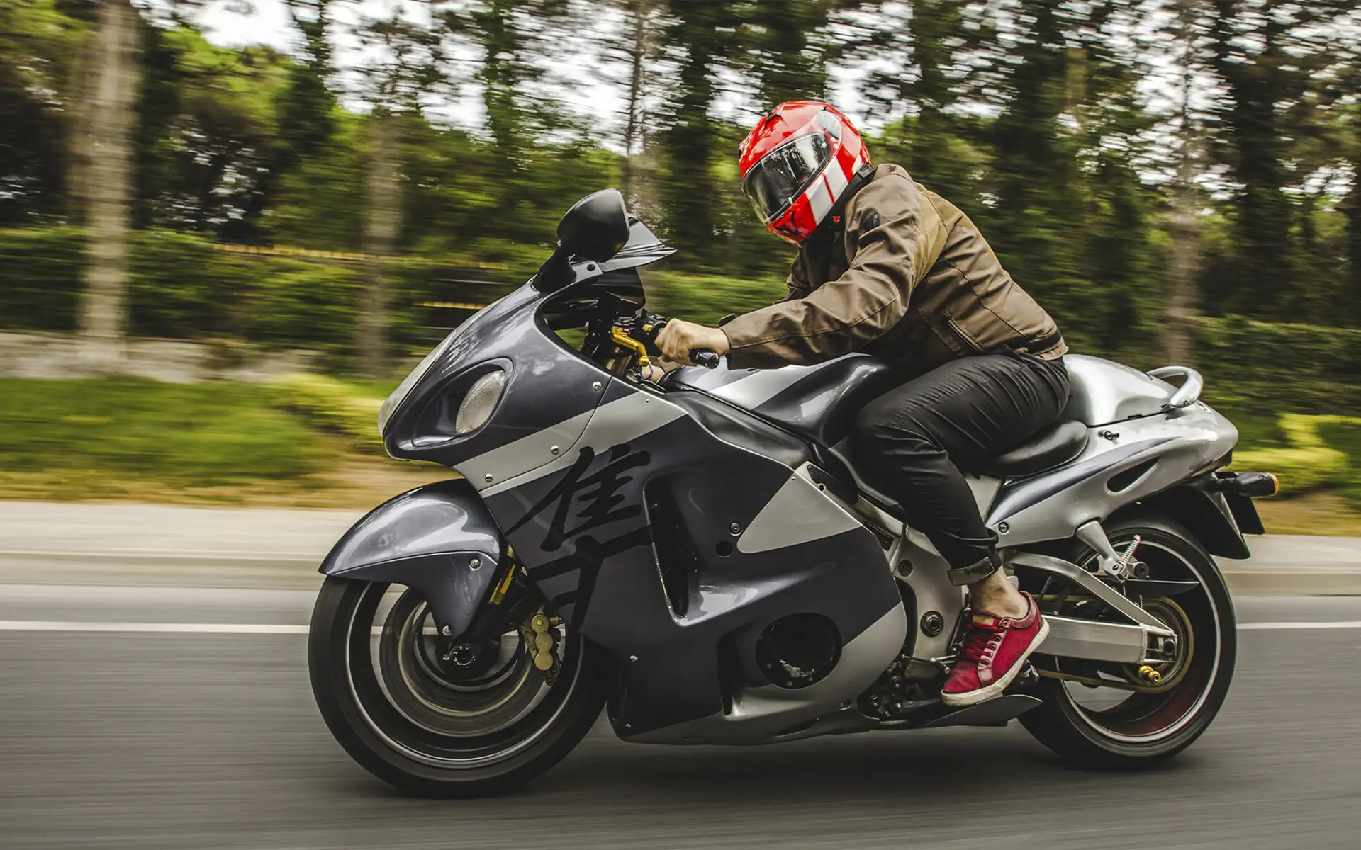 Fotografia de ação lateral de um motociclista com capacete vermelho a pilotar uma Suzuki Hayabusa cinzenta prateada em alta velocidade numa estrada ladeada por árvores desfocadas.