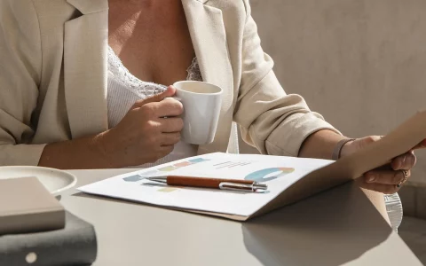 Plano focado em profissional de blazer bege a segurar uma caneca de café e a analisar relatórios com gráficos coloridos impressos sobre a mesa.