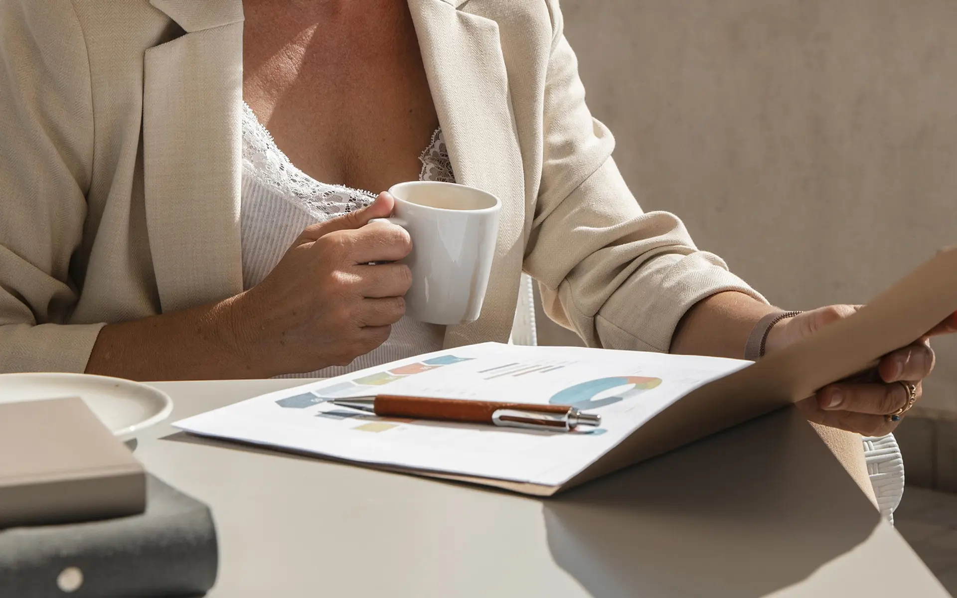 Plano focado em profissional de blazer bege a segurar uma caneca de café e a analisar relatórios com gráficos coloridos impressos sobre a mesa.