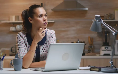 Jovem com blusa às bolinhas e cabelo apanhado sentada de forma pensativa em frente ao computador portátil, numa mesa iluminada por um candeeiro de secretária com a cozinha ao fundo.