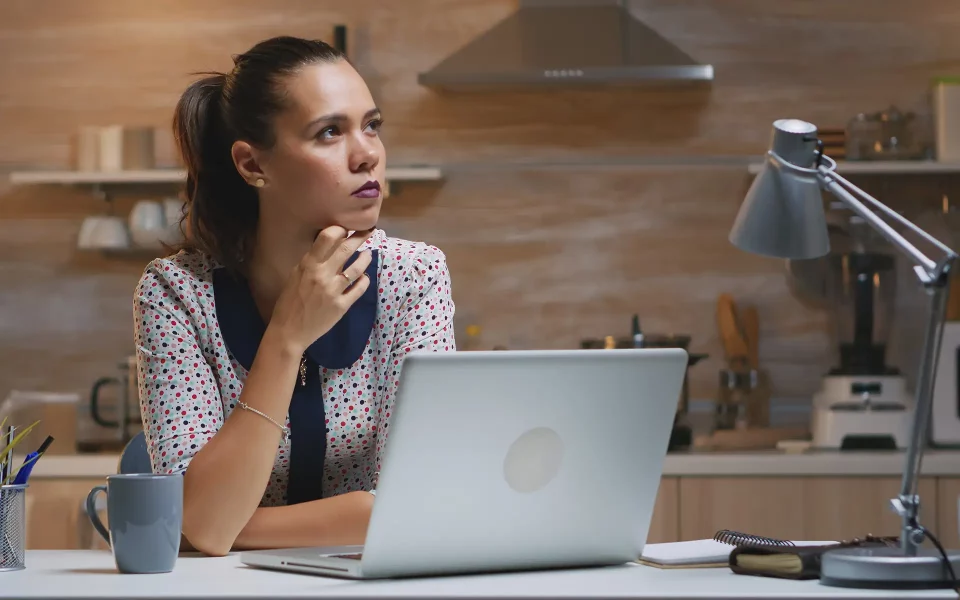 Jovem com blusa às bolinhas e cabelo apanhado sentada de forma pensativa em frente ao computador portátil, numa mesa iluminada por um candeeiro de secretária com a cozinha ao fundo.