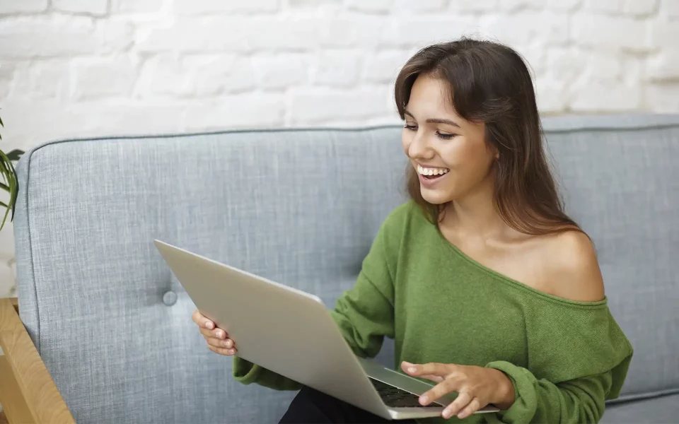 Jovem sorridente com camisola verde sentada no sofá a olhar e a reagir com alegria ao ecrã do portátil.