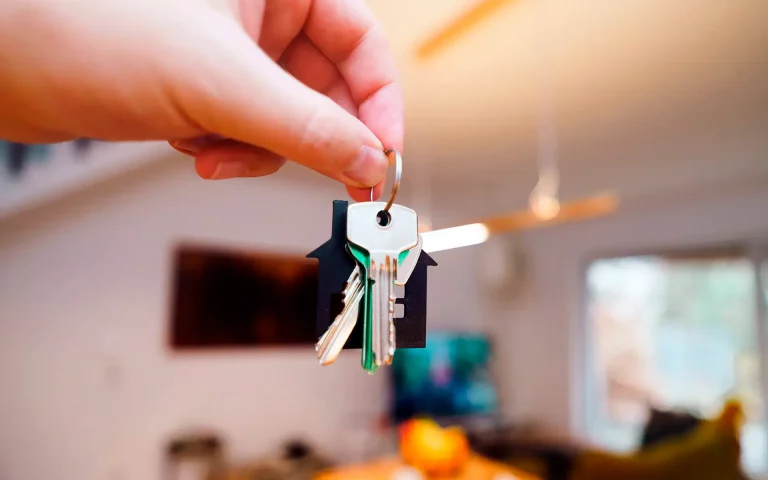 Plano focado em mão segurando um molho de chaves com um chaveiro em formato de casa preta, com um interior doméstico desfocado ao fundo.
