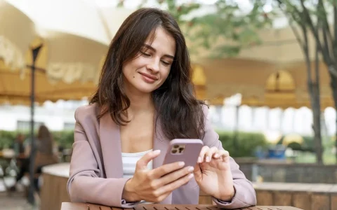 Jovem com blazer rosa velho sentada numa esplanada a sorrir enquanto consulta o seu smartphone.