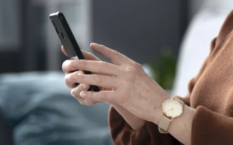 Plano aproximado das mãos de uma pessoa sénior a utilizar um smartphone preto, usando uma camisola de malha castanha e um relógio analógico com bracelete bege.