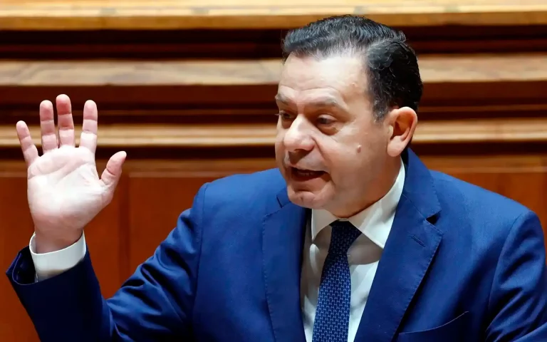 Luís Montenegro em fato azul a discursar com a mão direita erguida num ambiente parlamentar de madeira