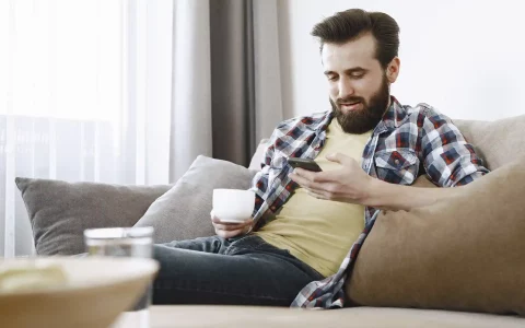 Organizar as financas no telemovel Homem com barba sentado num sofá a consultar o telemóvel enquanto segura uma chávena branca numa sala iluminada.