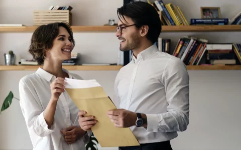 Casal jovem com camisas brancas a sorrir enquanto segura um envelope pardo com documentos num espaço luminoso