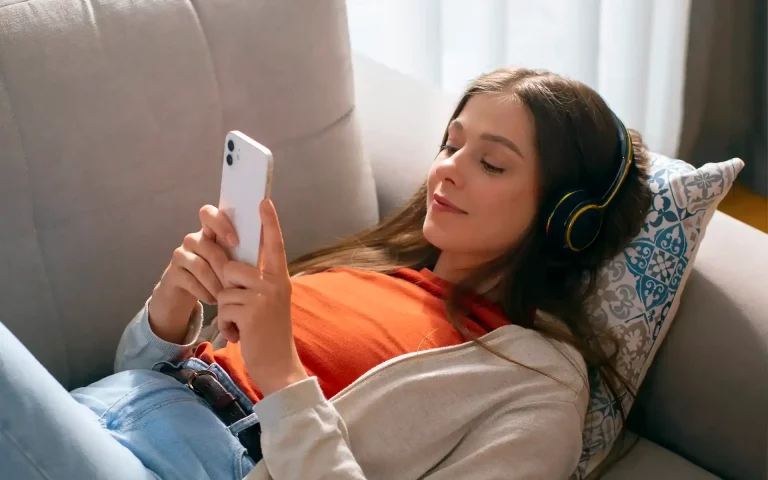 Jovem reclinada num sofá a usar auscultadores e a olhar para o smartphone branco que segura nas mãos.