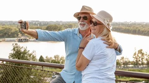 reforma antecipada Casal sénior com chapéus de palha e óculos de sol a tirar uma selfie no telemóvel num miradouro junto a um rio, a aproveitar a reforma antecipada.