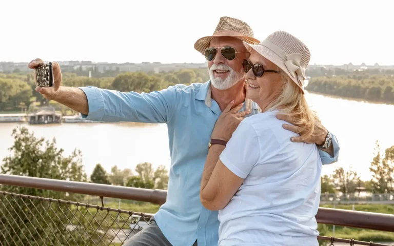 Casal sénior com chapéus de palha e óculos de sol a tirar uma selfie no telemóvel num miradouro junto a um rio, a aproveitar a reforma antecipada.